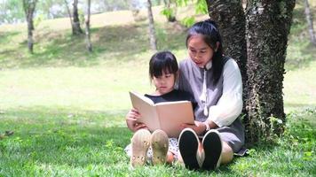 la madre e la figlia asiatiche felici si siedono sotto un albero sul prato del giardino estivo e leggono un libro insieme. video