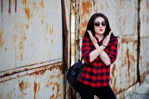 chica de retrato de moda con labios rojos con una camisa a cuadros roja, gafas de sol y una cerca oxidada de fondo de mochila. foto