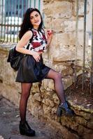 Young goth girl on black leather skirt and high heels punk shoes with backpack posed against iron fence. photo