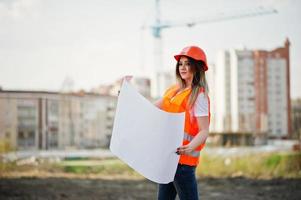 mujer ingeniera constructora con chaleco uniforme y casco protector naranja sostiene papel comercial contra nuevos edificios con grúa. foto