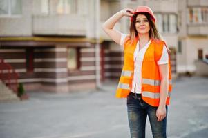 Engineer builder woman in uniform waistcoat and orange protective helmet against new building. Property living block theme. photo