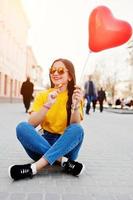 joven adolescente sentada en las calles de la ciudad con piruleta y globo de corazón en las manos, vestida con camiseta amarilla, jeans y gafas de sol. foto