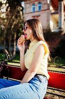 Young teenage girl sitting on bench with ice cream at hands, wear on yellow t-shirt, jeans and sunglasses. photo