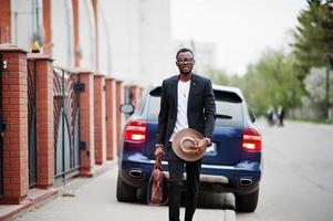 Stylish black man at glasses with hat, wear on suit with handbag against luxury car. Rich african american businessman. photo