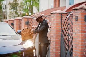 Stylish black man at glasses with hat, wear on suit with handbag against luxury car. Rich african american businessman. Look at watches. photo