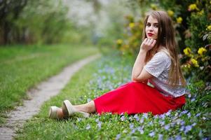 retrato de una hermosa chica sentada con labios rojos en el jardín de flores de primavera sobre césped con flores, vestido rojo y blusa blanca. foto