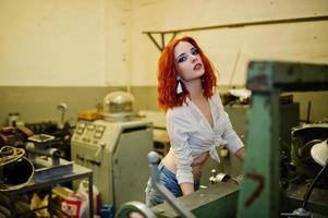 Red haired girl wear on short denim shorts and white blouse posed at industrial machine at the factory. photo