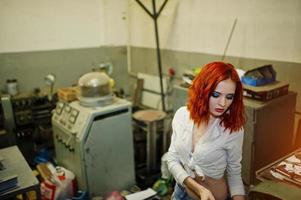 Red haired girl wear on short denim shorts and white blouse posed at industrial machine at the factory. photo