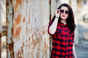 Fashion portrait girl with red lips wearing a red checkered shirt and sunglasses background rusty fence.