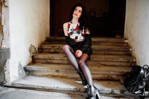 Young goth girl on black leather skirt and jacket with backpack posed on stairs of old house. photo