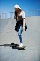 Young teenage urban girl with skateboard, wear on glasses, cap and ripped jeans at skate park on the evening. photo