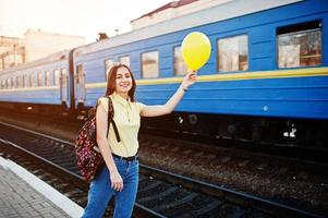 joven adolescente de pie en el andén de la estación de tren con globo a mano, vestida con camiseta amarilla, jeans y gafas de sol, con mochila. foto