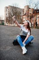 Young teenage urban girl with skateboard, wear on glasses, cap and ripped jeans at the yard sports ground on sunset making selfie on phone.
