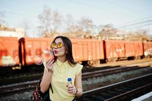 joven adolescente de pie en el andén de la estación de tren y soplando burbujas de jabón, vestida con camiseta amarilla, jeans y gafas de sol, con mochila.