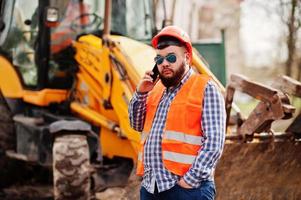 Brutal beard worker man suit construction worker in safety orange helmet, sunglasses against traktor with mobile phone at hand.