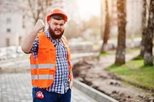 Portrait of brutal angry beard worker man suit construction worker in safety orange helmet against pavement with showing arms. photo