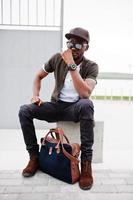 retrato de un elegante hombre afroamericano sentado con gafas de sol y gorra al aire libre. hombre negro de moda callejera. foto