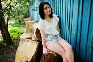 Portrait of sexy brunette girl on women's jeans shorts and white blouse against blue wooden house with stumps. photo