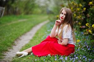 Portrait of sitiing beautiful girl with red lips at spring blossom garden on grass with flowers, wear on red dress and white blouse. photo