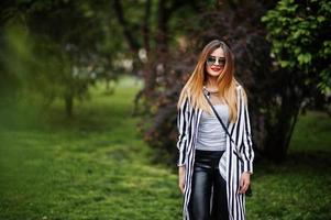 Fashionable woman look with black and white striped suit jacket, leather pants and sunglasses posing against bushes at street. Concept of fashion girl. photo