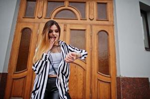 Fashionable woman look with black and white striped suit jacket, leather pants, posing against large wooden door of building and looking on watches. Concept she is late. photo