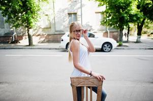 Stylish blonde woman wear at jeans, glasses, choker and white shirt near chair against luxury car. Fashion urban model portrait. photo