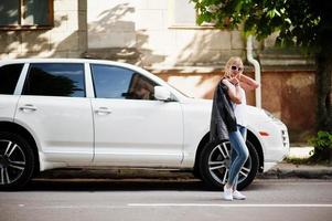elegante mujer rubia vestida con jeans, gafas de sol y camisa blanca contra un auto de lujo. retrato de modelo urbano de moda.