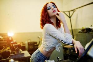 Red haired girl wear on short denim shorts and white blouse posed at industrial machine at the factory. photo