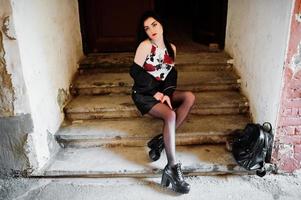 Young goth girl on black leather skirt and jacket with backpack posed on stairs of old house. photo