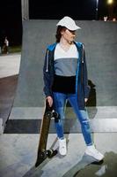 Young teenage urban girl with skateboard, wear on glasses, cap and ripped jeans at skate park on the evening. photo