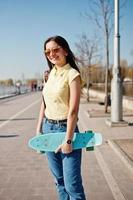 Young teenage girl with small skateboard, penny board, wear on yellow t-shirt, jeans and sunglasses. photo