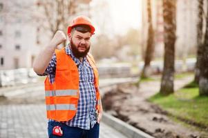 Portrait of angry beard worker man suit construction worker in safety orange helmet against pavement with showing arms.