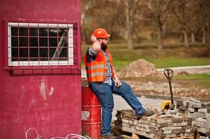 trabajador de barba brutal traje de hombre trabajador de la construcción en casco naranja de seguridad sentado en barril rojo. foto