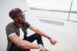 retrato de un elegante hombre afroamericano sentado con gafas de sol y gorra con bolso al aire libre. hombre negro de moda callejera. foto