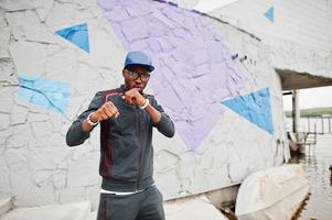 Portrait of stylish african american man on sportswear, cap and glasses against colored wall. Black men model street fashion. Punch boxer. photo