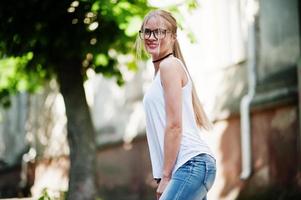 Stylish blonde woman wear at jeans, glasses, choker and white shirt. Fashion urban model portrait. photo