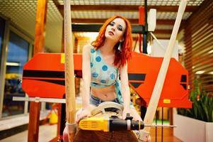 Red haired model sitting on wood sawmill with drill at store or household shop of working tools. photo