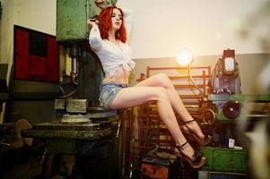 Red haired girl wear on short denim shorts and white blouse posed at industrial machine at the factory. photo