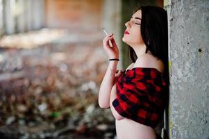 Retrato de una chica fumadora con labios rojos que llevaba una camisa a cuadros roja con los hombros desnudos posó un lugar sexy de fondo abandonado. foto