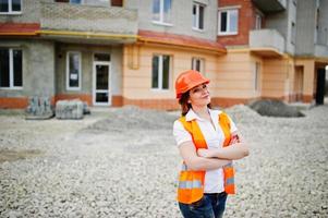 Engineer builder woman in uniform waistcoat and orange protective helmet against new building. Property living block theme. photo