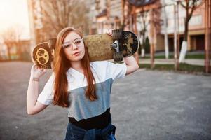 joven adolescente urbana con monopatín, gafas, gorra y jeans rotos en el campo de deportes del patio al atardecer. foto