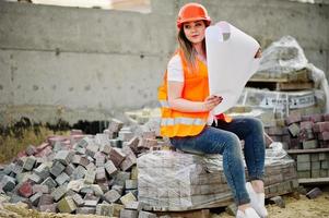 Engineer builder woman in uniform waistcoat and orange protective helmet hold business layout plan paper sitting on pavement.