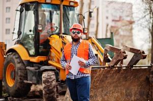 Brutal beard worker man suit construction worker in safety orange helmet, sunglasses against traktor with plan paper at hands. photo