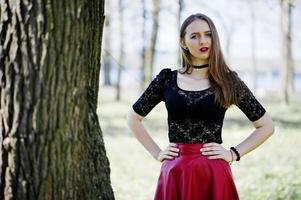 retrato de niña con maquillaje brillante con labios rojos, collar de gargantilla negra en el cuello y falda de cuero rojo en el parque de primavera. foto