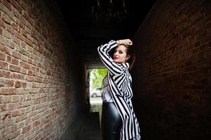 espalda de mujer de moda con chaqueta de traje a rayas blancas y negras, pantalones de cuero, posando en el antiguo túnel de ladrillo rojo de la calle. concepto de chica de moda. foto