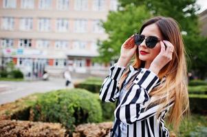Close up portrait of fashionable woman look with black and white striped suit jacket and sunglasses posing against bushes at street. Concept of fashion girl. photo