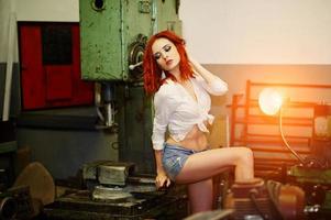 Red haired girl wear on short denim shorts and white blouse posed at industrial machine at the factory. photo