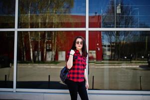 Fashion portrait girl with red lips wearing a red checkered shirt and backpack with sunglasses background large mirror windows. photo