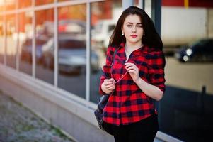 Fashion portrait girl with red lips wearing a red checkered shirt and backpack holding sunglasses at hands background large mirror windows. photo