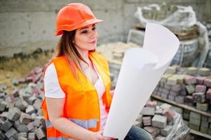 mujer ingeniera constructora con chaleco uniforme y casco protector naranja sostiene papel de plan de diseño de negocios sentado en el pavimento.